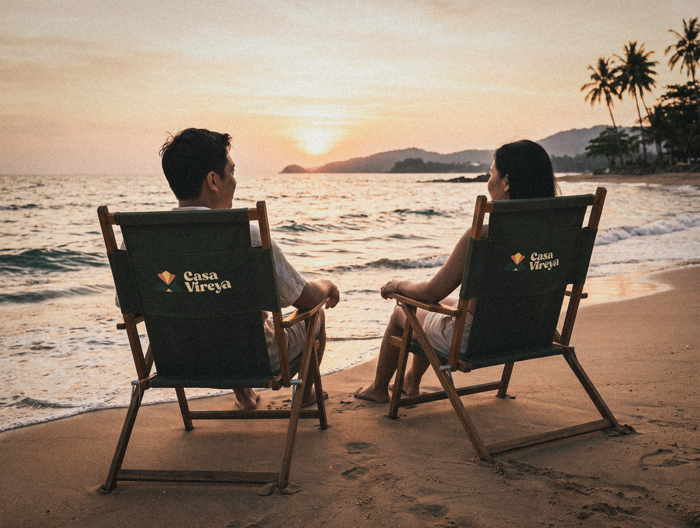 A couple sits in weathered canvas chairs at the water's edge, watching the sun lower behind distant hills.