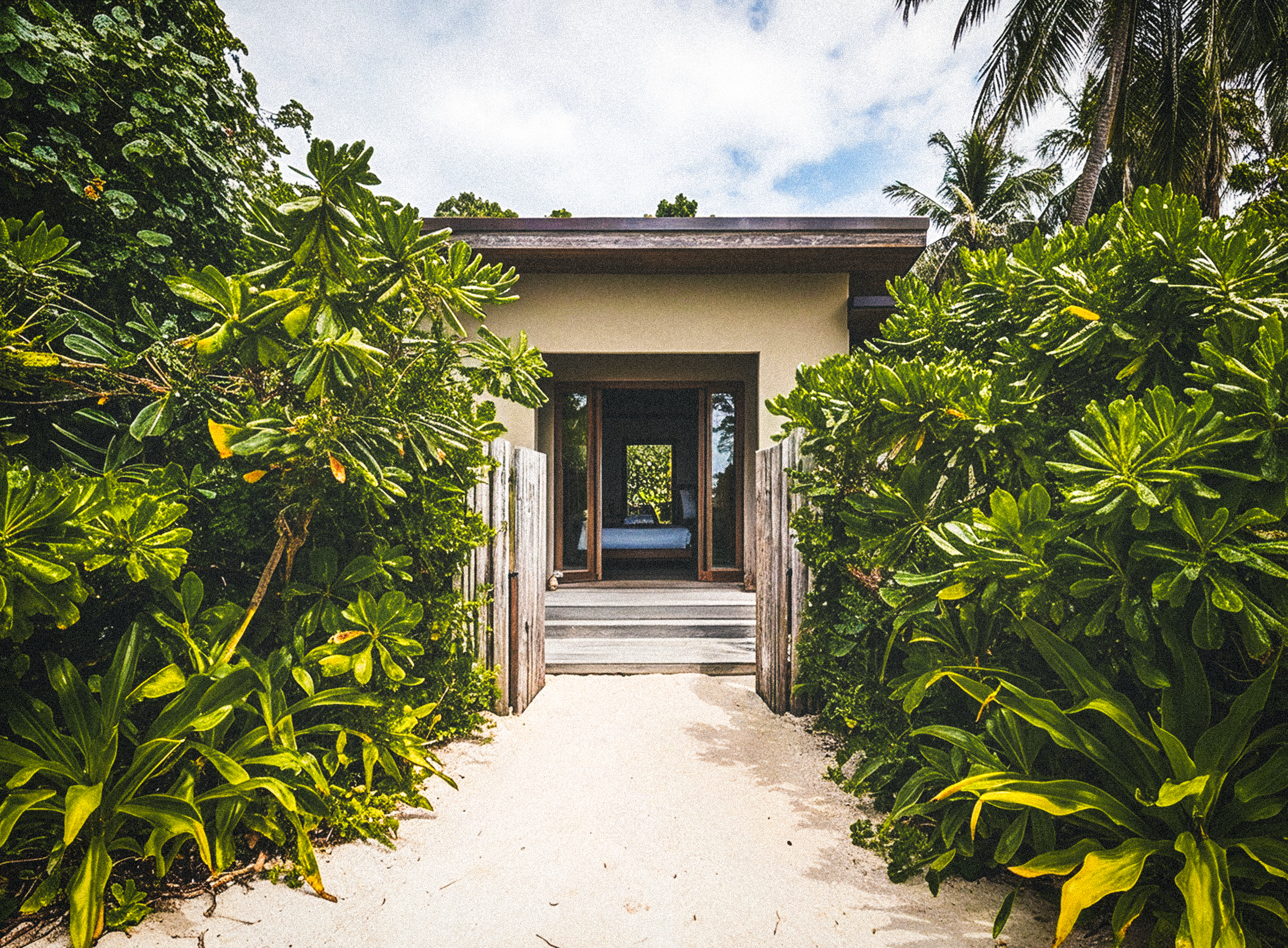 A sandy path lined with lush green shrubs leading toward the villa's open bedroom doors.