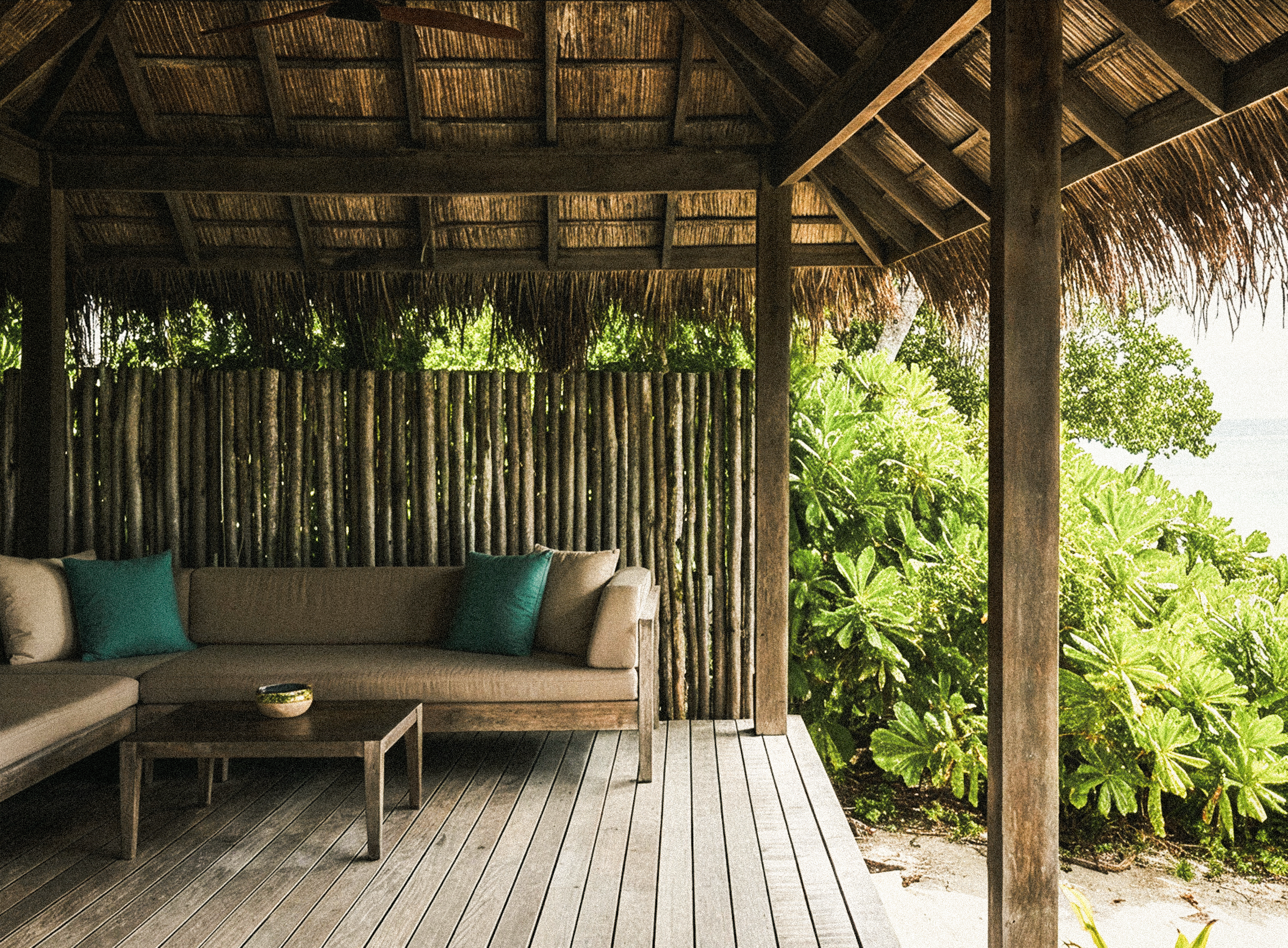 A covered outdoor lounge with a low sofa and teal cushions, overlooking the greenery toward the sea.