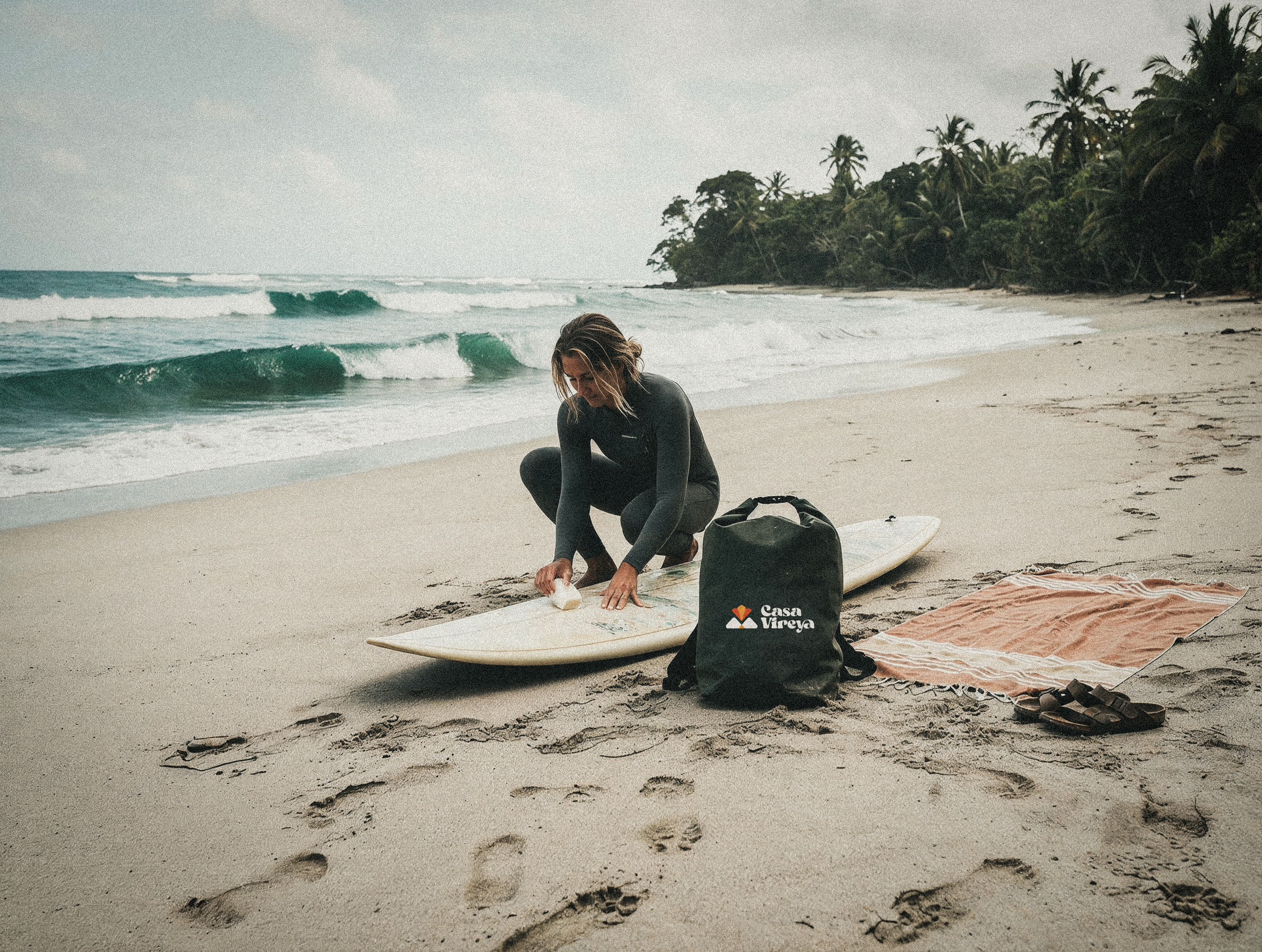 A surfer kneels beside her board on the shoreline, waxing it before paddling out toward the break.