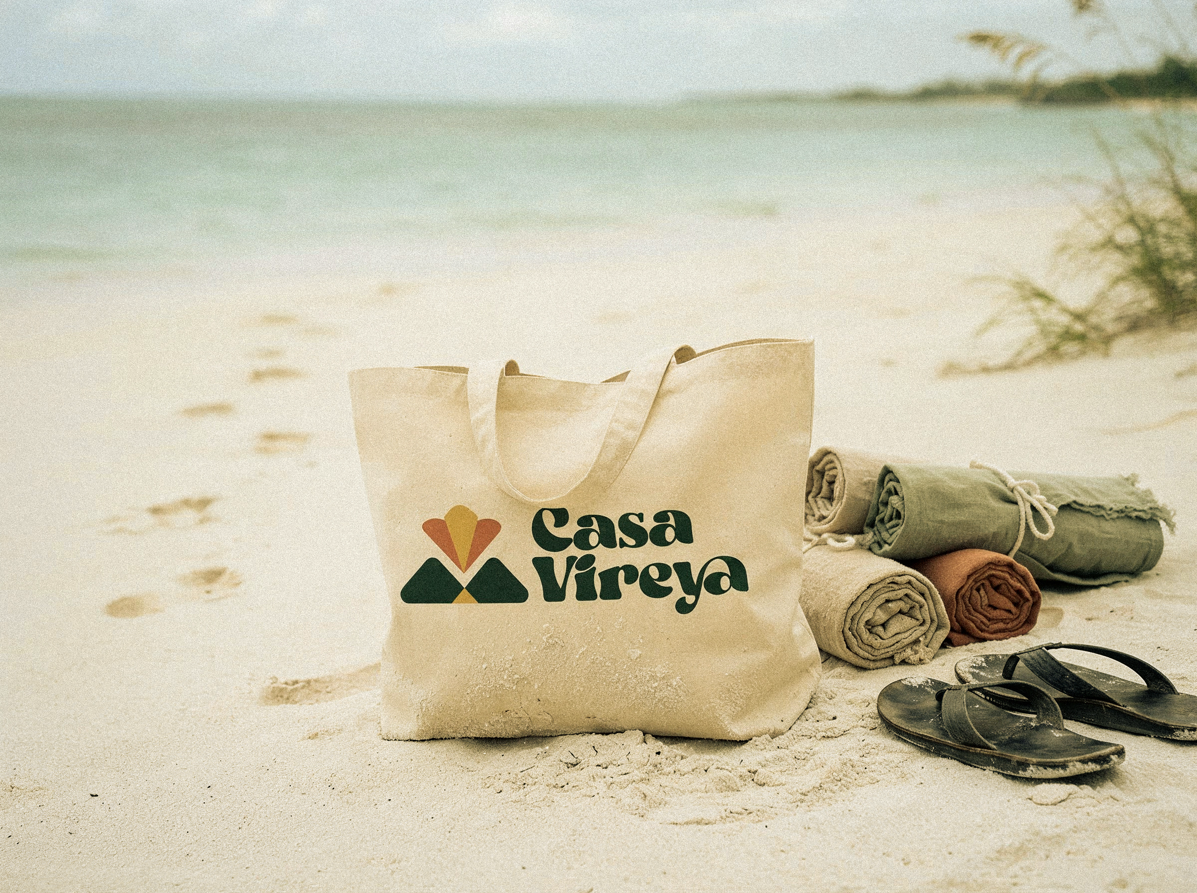 A canvas Casa Vireya tote bag resting on the sand beside rolled linen towels.